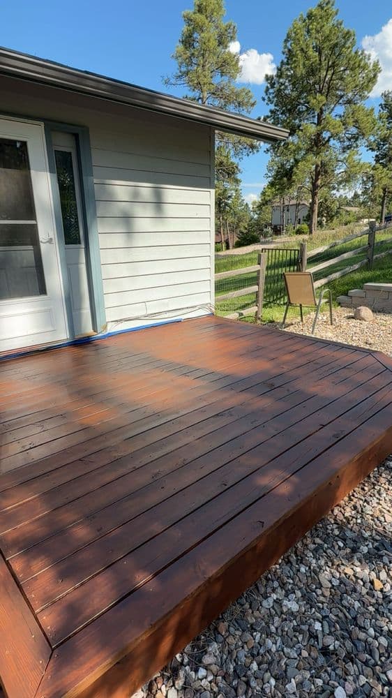 Ground-level deck freshly stained a deep red-brown, still wet-looking in the sun, against pale siding