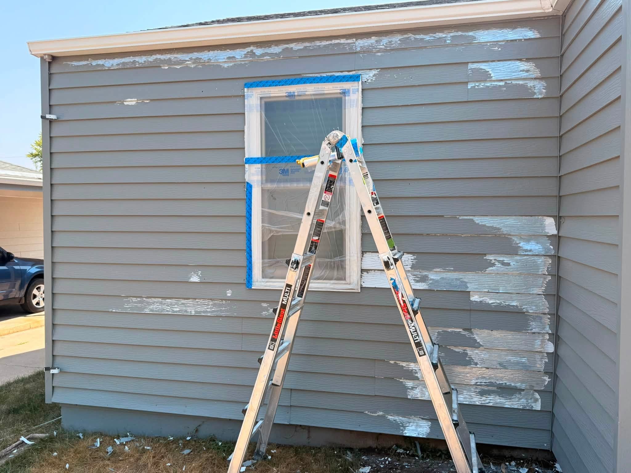 Lap siding before the new coating: the old paint gone in wide patches with a pale layer showing underneath, one window masked off with tape and film, an A-frame ladder standing in front of the wall and scraped chips on the ground